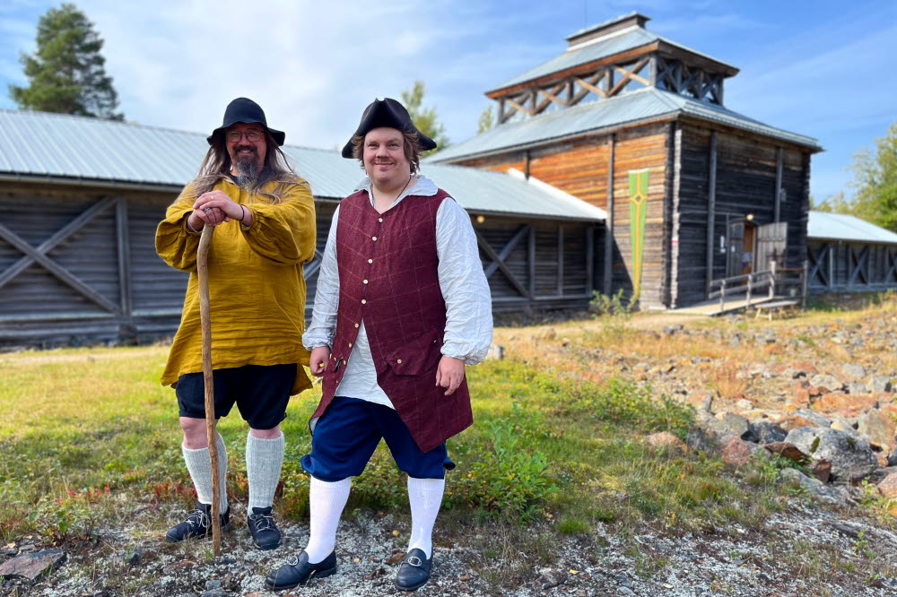 Två män i helfigur klädda i 1700-talskläder står på stenig mark framför en träbyggnad i solsken.