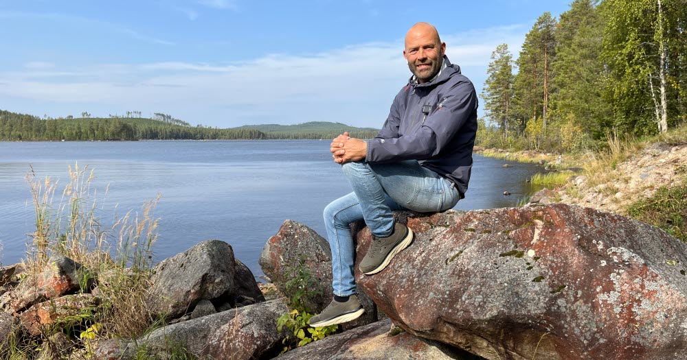 En person i helfigur sitter på sten framför en blå sjö. Sommar.