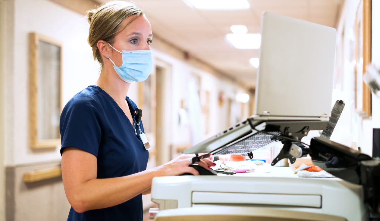 Registered nurse wearing a face mask documents medicine provided to a patient into a laptop computer at the nursing station at work, Indiana, USA