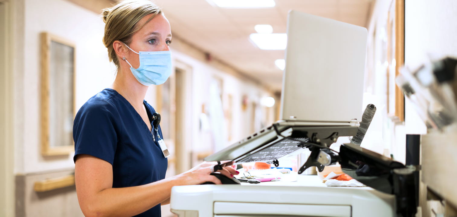 Registered nurse wearing a face mask documents medicine provided to a patient into a laptop computer at the nursing station at work, Indiana, USA