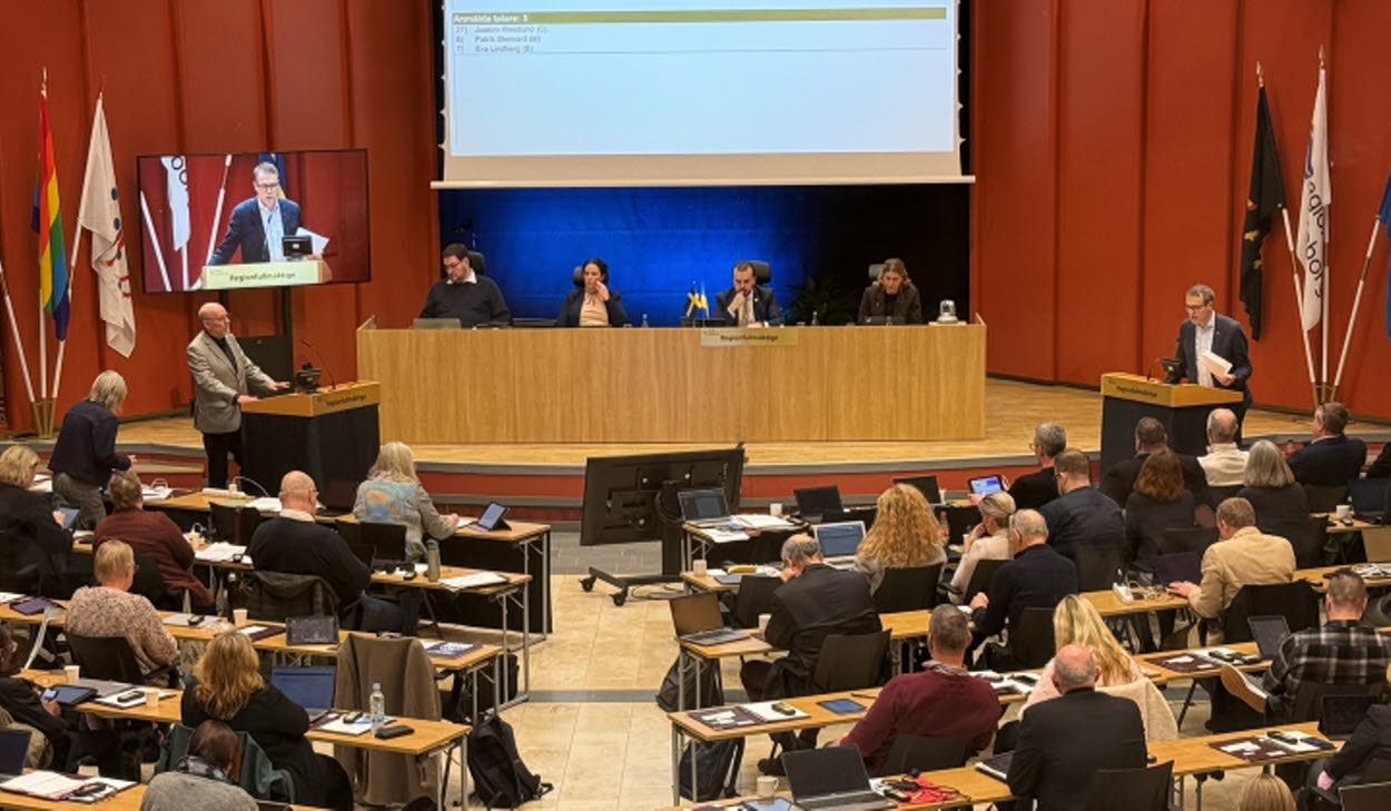 En fullsatt regionfullmäktigesal med deltagare vid bärbara datorer och dokument på borden. En person talar vid en talarstol, och en presentation visas på en stor skärm med text och en talartidräknare. Jan Lahenkorva och Patrik Stenvard debatterar.