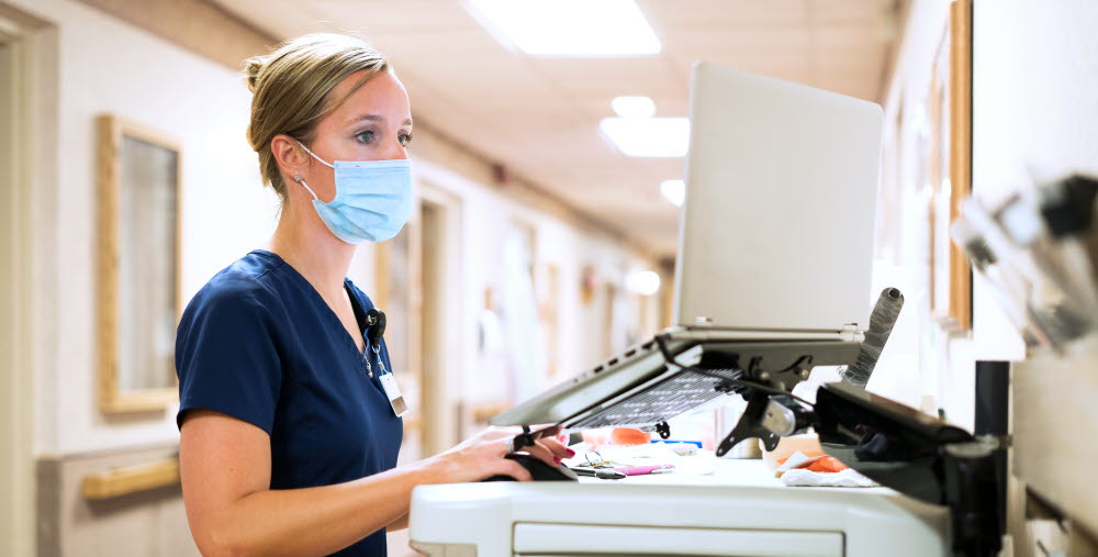 Registered nurse wearing a face mask documents medicine provided to a patient into a laptop computer at the nursing station at work, Indiana, USA