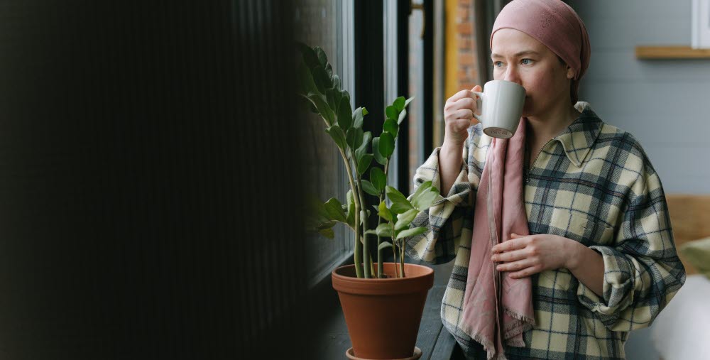 Person står vid ett fönster och dricker ur en kopp. I fönstret står en krukväxt.